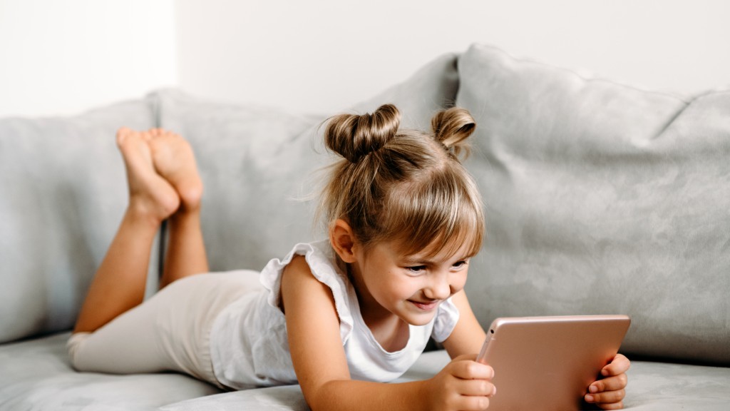 Menina pequena deitada em um sofá cinza sorrindo enquanto assiste ao Luccas Toon no tablet.