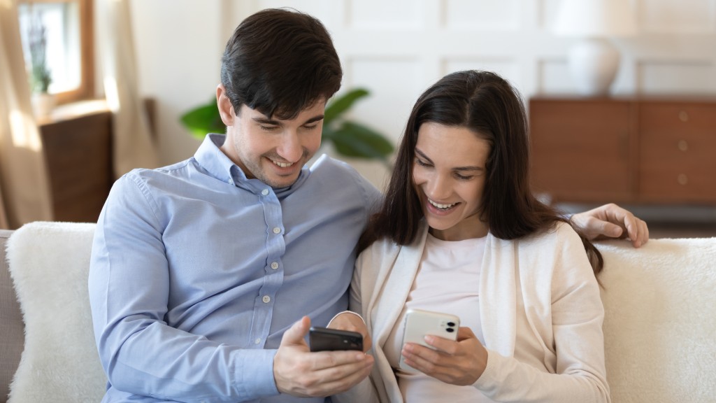Casal sorridente sentado no sofá, ele de camisa azul e ela de blusa branca, cada um segurando seu celular enquanto realizam a transferência de contatos de um celular Android para o iPhone.