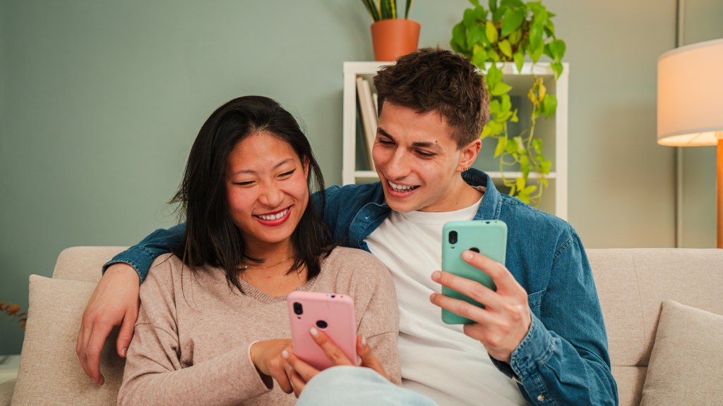 Casal sorridente sentado no sofá, cada um segurando seu celular, representando a transferência de contatos de um celular para o outro.