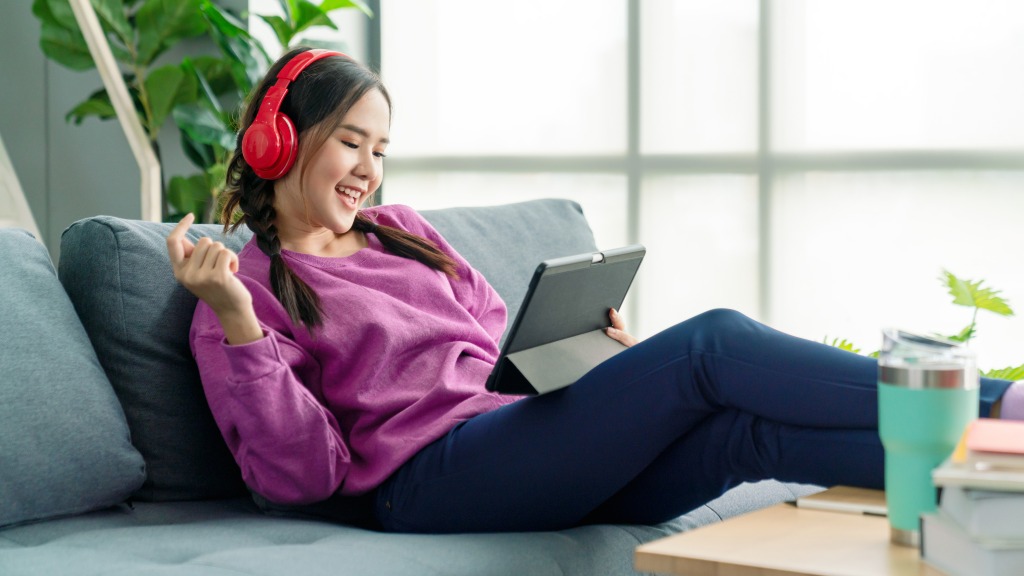Garota adolescente com blusa rosa e headphones vermelhos, feliz, sentada no sofá enquanto usa o tablet para navegar online, representando uso de internet em Ribeirão Preto.