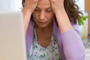 Mulher de blusa roxa sentada à mesa em frente ao notebook, com expressão de frustração após sofrer golpe na internet.