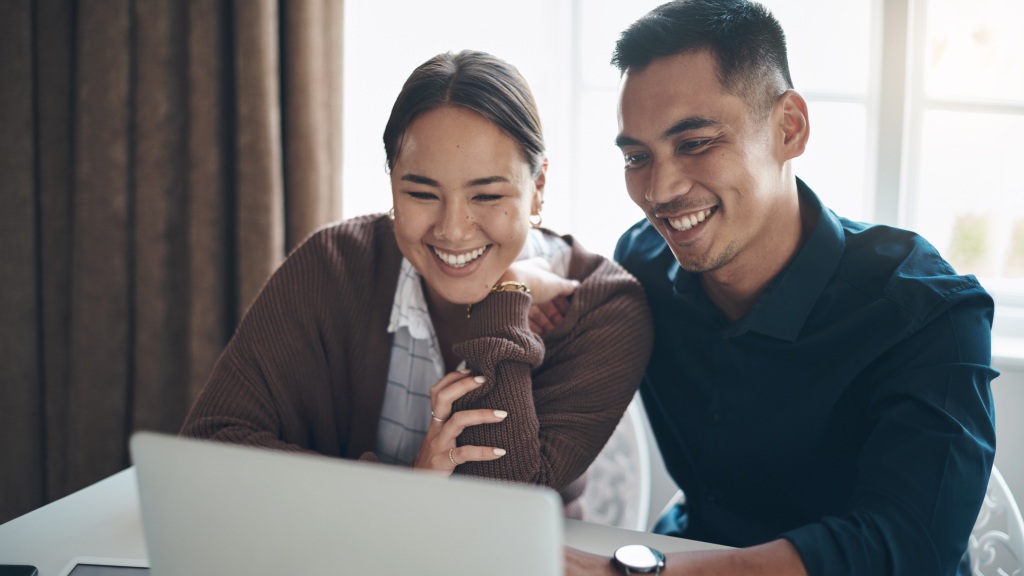 Casal sorridente, ele usa camisa azul escura e ela blusa marrom, sentados na mesa enquanto usam o notebook para navegar online, representando o uso de internet em Ribeirão Preto.