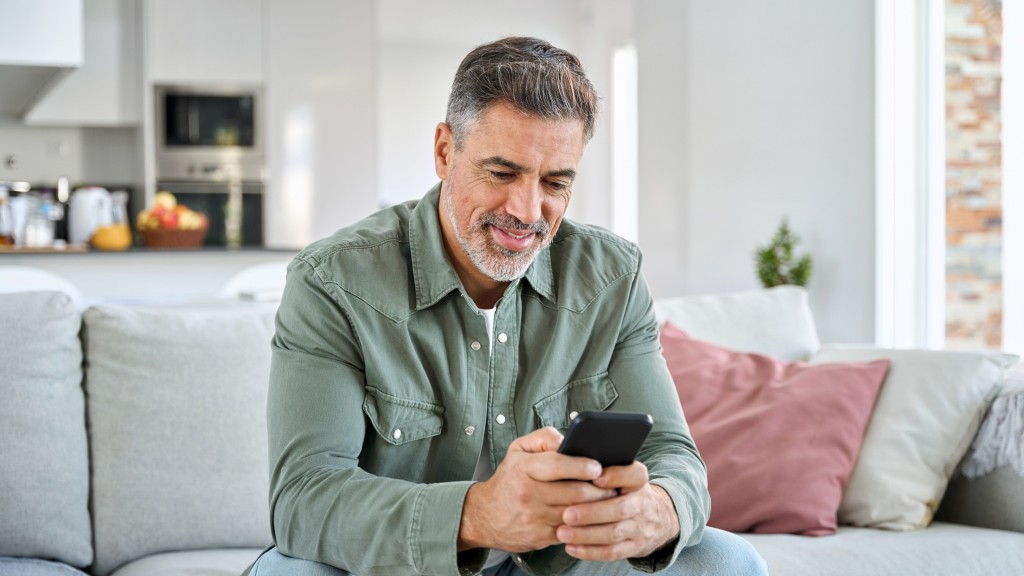 Homem de meia idade vestindo camisa verde sentado em um sofá cinza enquanto configura mensagem automática no WhatsApp pelo celular.
