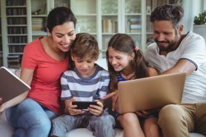 Família com pai, mãe e dois filhos, um menino e uma menina, sentados no sofá, sorridentes, enquanto navegam na internet pelo notebook e celular.