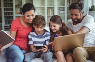Família com pai, mãe e dois filhos, um menino e uma menina, sentados no sofá, sorridentes, enquanto navegam na internet pelo notebook e celular.