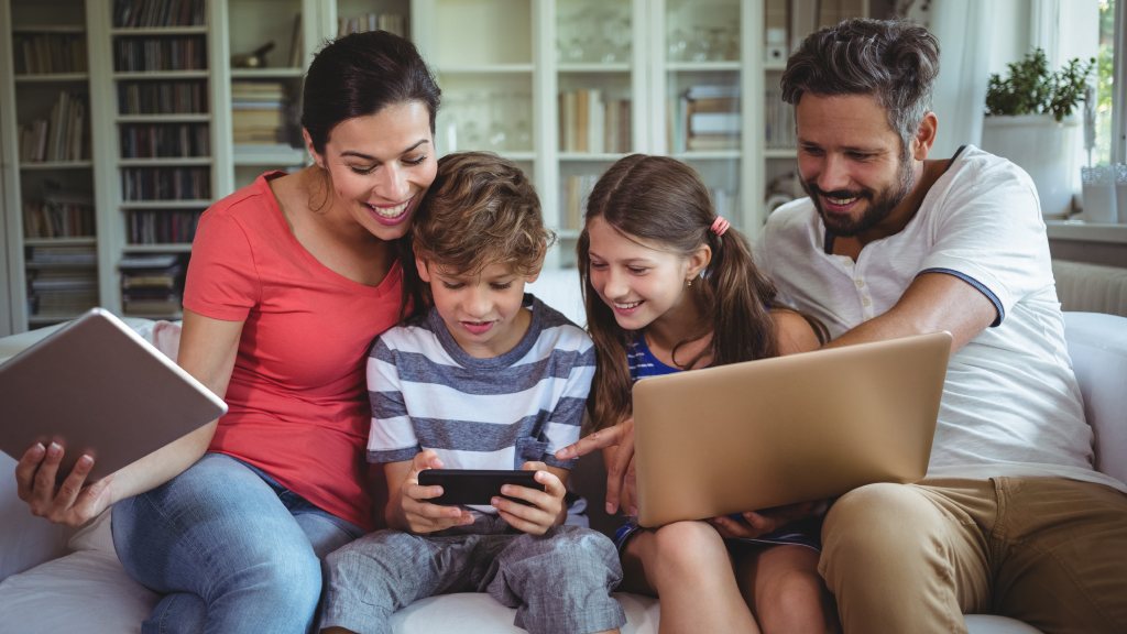 Família com pai, mãe e dois filhos, um menino e uma menina, sentados no sofá, sorridentes, enquanto navegam na internet pelo notebook e celular.
