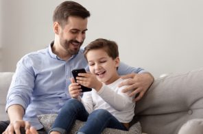 Pai e filho sorrindo no sofá enquanto o filho assiste conteúdos do Luccas Toon no celular, com o pai ao lado olhando e acompanhando a brincadeira.