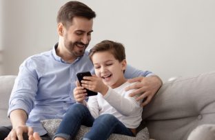 Pai e filho sorrindo no sofá enquanto o filho assiste conteúdos do Luccas Toon no celular, com o pai ao lado olhando e acompanhando a brincadeira.