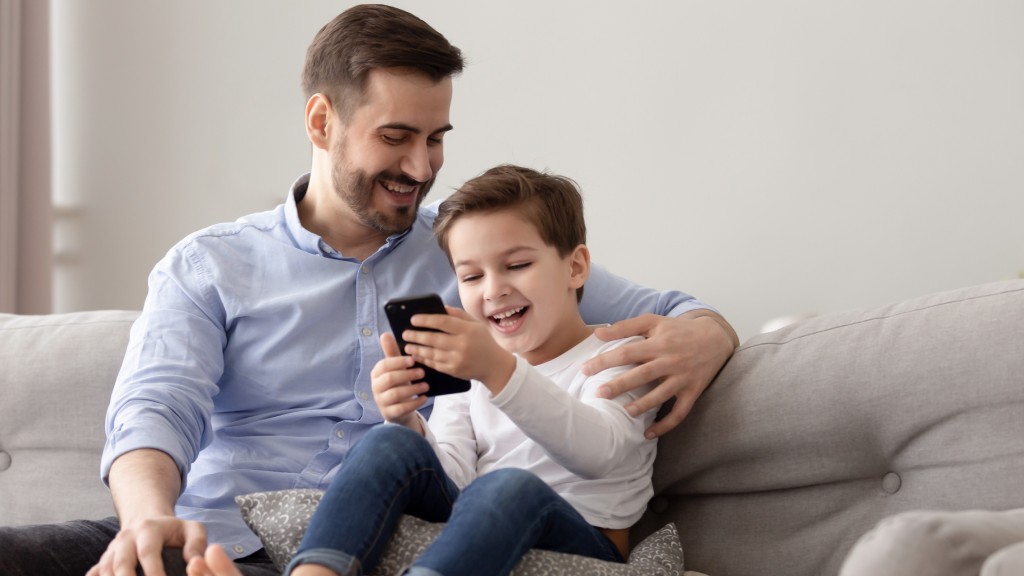 Pai e filho sorrindo no sofá enquanto o filho assiste conteúdos do Luccas Toon no celular, com o pai ao lado olhando e acompanhando a brincadeira.