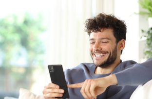 Homem jovem feliz com blusa moletom azul escuto navegando no celular conectado à rede Wi-Fi.
