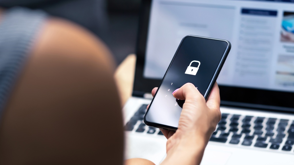 Pessoa segurando um celular com a tela de bloqueio exigindo senha, enquanto um notebook está sobre a mesa ao fundo.