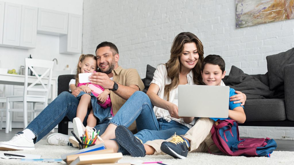 Família conectada em dispositivos Wi-Fi sentada no chão da sala; pai com filha no colo assistindo a conteúdo no celular e mãe com filho ao lado navegando juntos no notebook.
