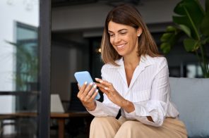 Mulher adulta vestindo camisa branca com linhas pretas e calça marrom, sentada em um sofá cinza enquanto coloca mensagem automática no WhatsApp pelo celular.