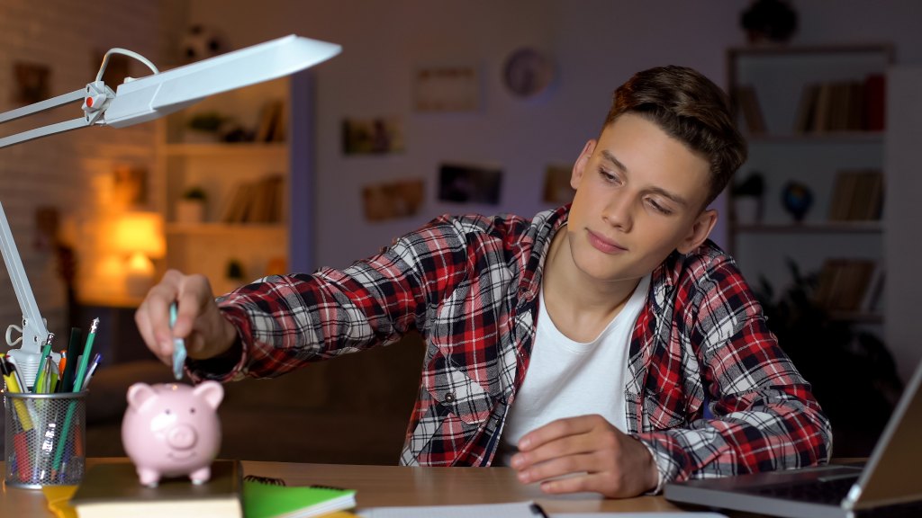 Adolescente de camisa xadrez e camiseta branca, sentado à escrivaninha do quarto, depositando nota em porquinho, simbolizando aprendizado financeiro com a Mooney, parceira da Desktop.