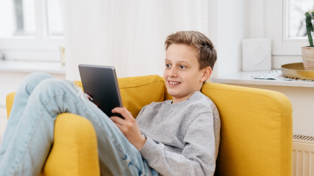 Menino de blusa cinza e calça jeans sorrindo, sentado no sofá enquanto lê HQs no tablet pelo app Desktop Comics.