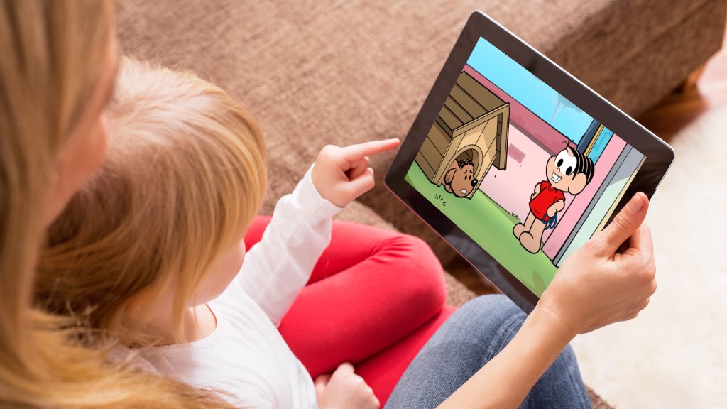Mãe e filha sentadas no sofá enquanto a mãe segura um tablet com um desenho da Turma da Mônica exibido pelo app Monicaverso.
