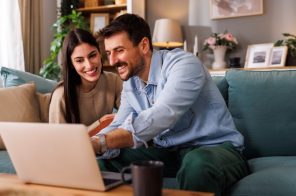 Casal sorridente sentado no sofá, usando notebook sobre a mesa enquanto pesquisa planos de internet para contratar na Black Friday.