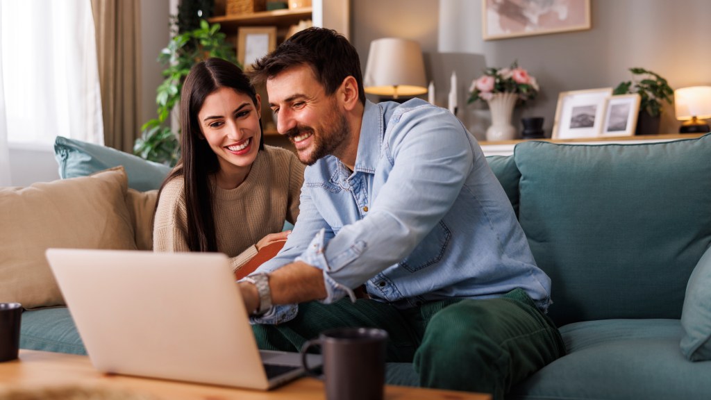 Casal sorridente sentado no sofá, usando notebook sobre a mesa enquanto pesquisa planos de internet para contratar na Black Friday.