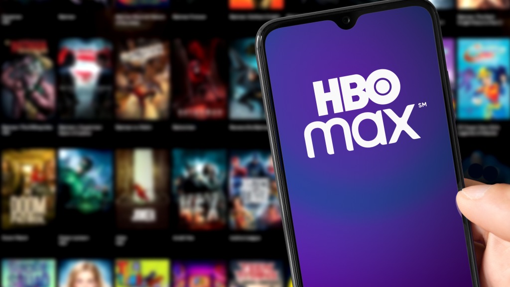 Mão segurando um celular com o app da HBO Max aberto, com projeção desfocada ao fundo de filmes e séries disponíveis no streaming.
