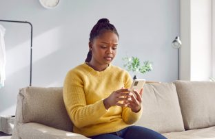 Mulher jovem de blusa amarela sentada no sofá da sala, segurando o celular com expressão de desconfiança diante de possíveis golpes no WhatsApp.