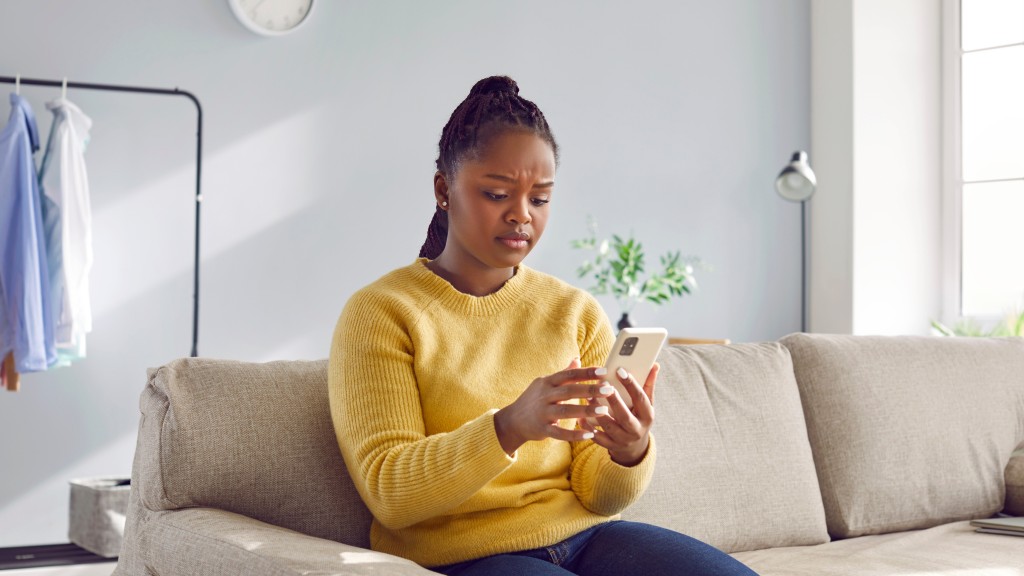 Mulher jovem de blusa amarela sentada no sofá da sala, segurando o celular com expressão de desconfiança diante de possíveis golpes no WhatsApp.