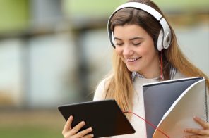 Jovem de blusa branca sorrindo, usando fones de ouvido brancos, segura um caderno e um tablet enquanto lê quadrinhos no app Desktop Comics.