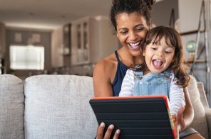 Mãe e filha sorridentes, sentadas no sofá cinza da sala de estar, segurando um tablet e felizes com a proteção parental ativa no dispositivo.