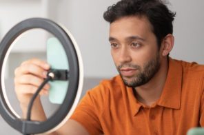 Alt text: Homem de camisa laranja sentado à mesa de trabalho com celular em suporte de ring light, utilizando o aparelho como webcam.