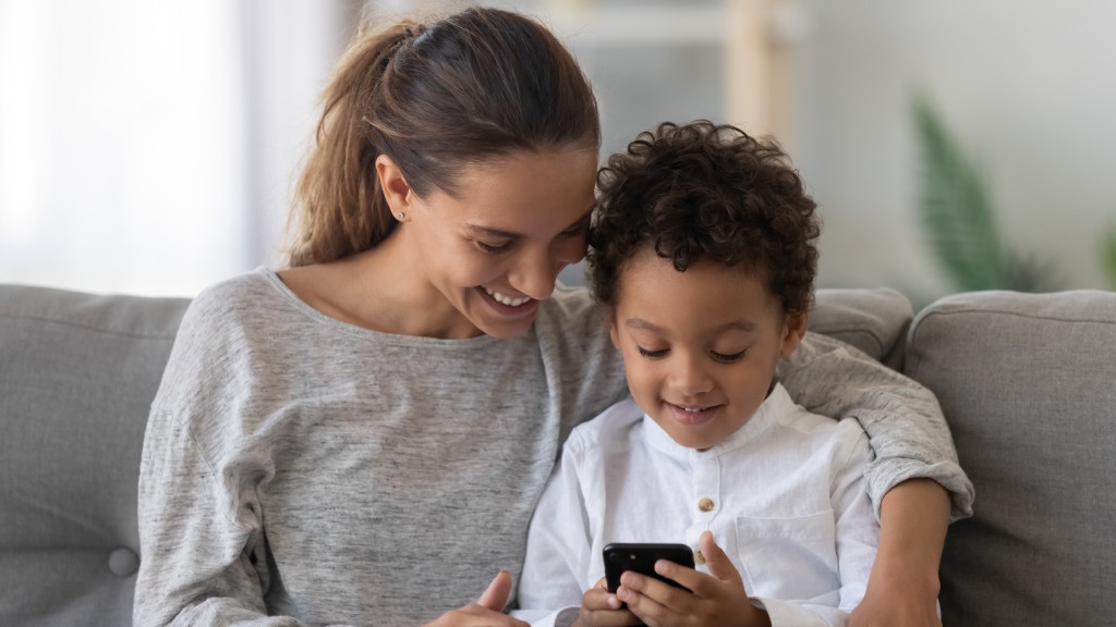 Mãe e filho sorridentes, ela de blusa cinza e ele de camisa branca, enquanto ele segura um celular e navega protegido com a proteção dos pais ativa no aparelho.
