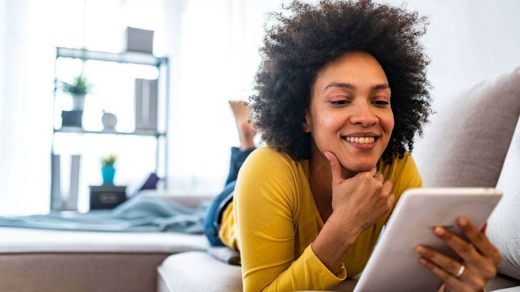 Mulher de blusa amarela e cabelos cacheados, sorrindo enquanto lê revistas no app Revistas Já deitada de bruços no sofá.