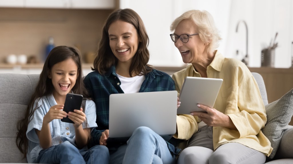 Criança mexendo no celular ao lado de mulher com tablet e mulher idosa com notebook, todas sorridentes, sentadas no sofá, representando segurança na internet para crianças e idosos.