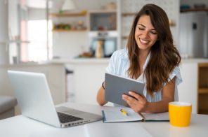 Mulher sentada à mesa com notebook, caderno, caneta e xícara, sorrindo com tablet na mão enquanto realiza download e upload com rapidez e qualidade.