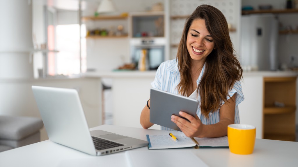 Mulher sentada à mesa com notebook, caderno, caneta e xícara, sorrindo com tablet na mão enquanto realiza download e upload com rapidez e qualidade.