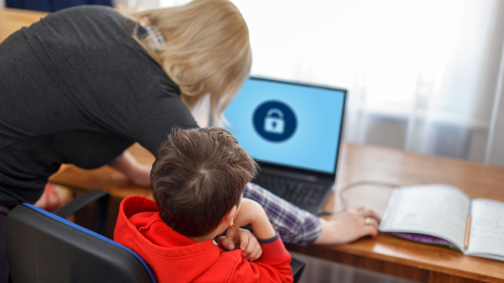 Criança sentada à mesa em frente a um notebook, enquanto a mãe, inclinada, define limites usando o controle dos pais. A tela mostra um cadeado e, ao lado do computador, há um caderno aberto.