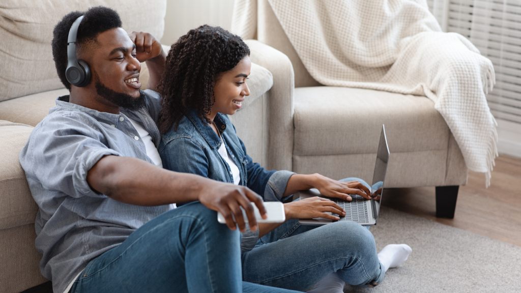 Casal sorridente sentado no chão da sala; ele usa headphones pretos para ouvir música, e ela está com as pernas cruzadas e um notebook no colo.