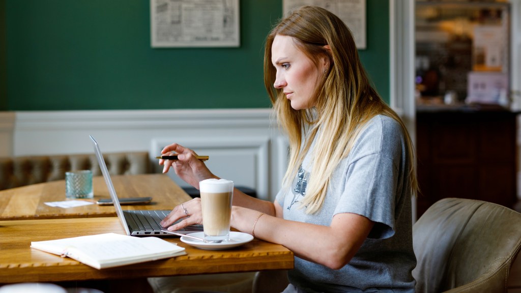 Moça jovem com cabelos loiros sentada em um café, com notebook em cima de mesa de madeira, ao lado de um copo de café, simbolizando o uso de Wi-Fi público e os cuidados necessários.