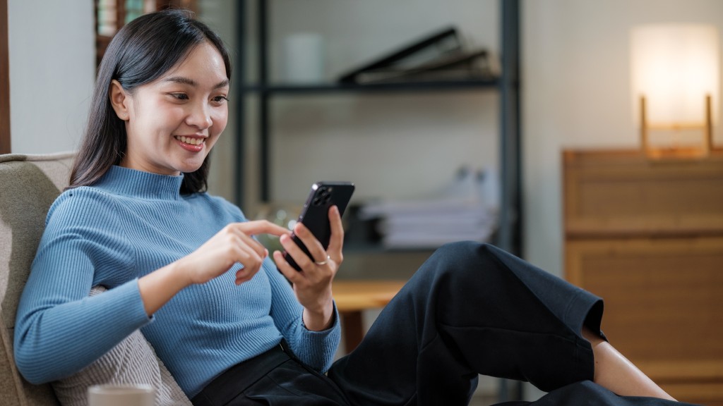 Mulher jovem oriental sorridente, de blusa azul e calça preta, sentada no sofá e segurando iPhone conectado a uma rede hotspot.