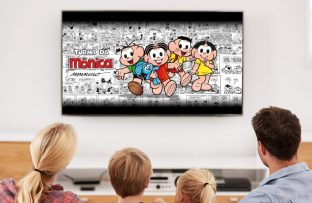 Pais com um menino e uma menina sentados em um sofá assistindo à Turma da Mônica na TV por meio do Monicaverso.