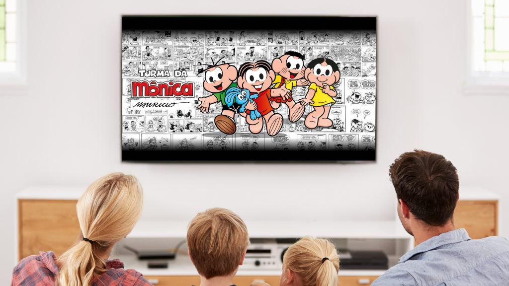 Pais com um menino e uma menina sentados em um sofá assistindo à Turma da Mônica na TV por meio do Monicaverso.