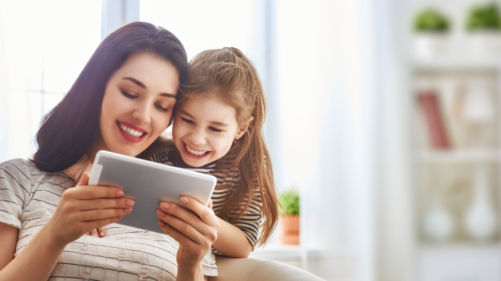 Mãe e filha sorridentes usando um tablet para acessar o Monicaverso; a mãe está sentada em uma poltrona e a filha abraça seu pescoço.