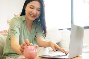 Moça oriental de verde, sentada no sofá em frente ao notebook na sala de estar, sorrindo enquanto deposita moeda em um porquinho sobre a mesa, simbolizando aprendizado financeiro com o app Mooney.