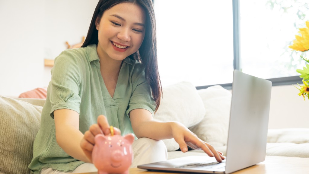 Moça oriental de verde, sentada no sofá em frente ao notebook na sala de estar, sorrindo enquanto deposita moeda em um porquinho sobre a mesa, simbolizando aprendizado financeiro com o app Mooney.