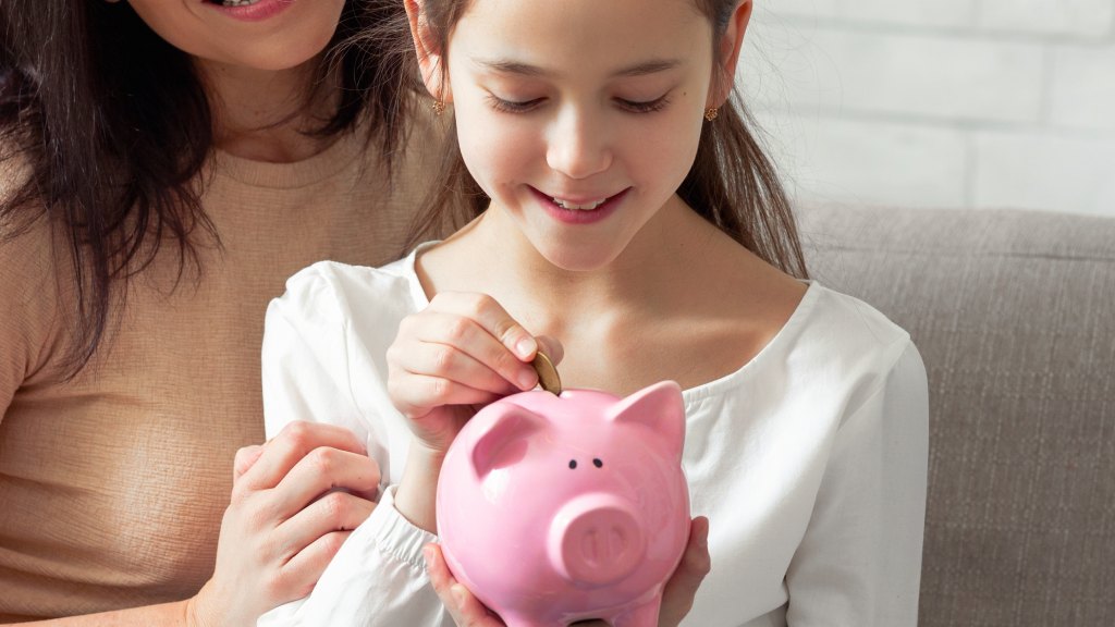 Criança de blusa branca sentada no sofá, sorrindo enquanto deposita moeda em porquinho rosa, com a mãe atrás, sorrindo e apoiando a mão na filha com carinho.