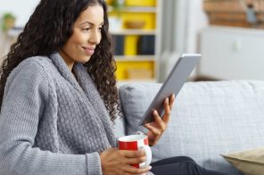 Mulher jovem de roupão cinza sentada no sofá, sorrindo enquanto lê conteúdos do app Revistas Já pelo tablet e segura uma caneca de porcelana.