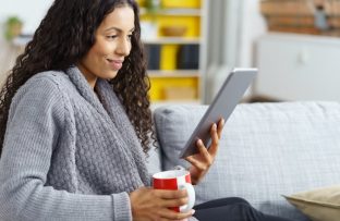 Mulher jovem de roupão cinza sentada no sofá, sorrindo enquanto lê conteúdos do app Revistas Já pelo tablet e segura uma caneca de porcelana.