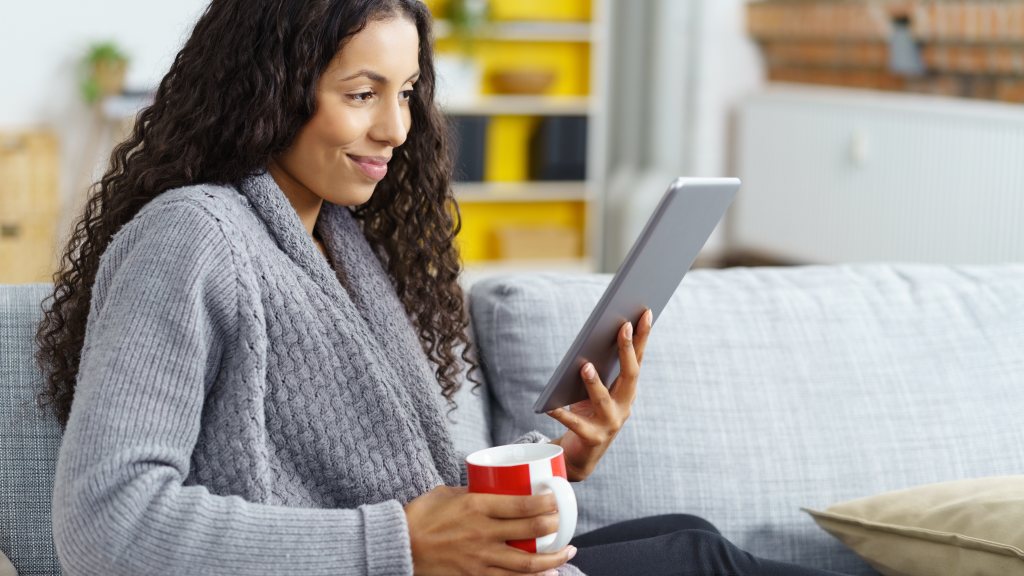 Mulher jovem de roupão cinza sentada no sofá, sorrindo enquanto lê conteúdos do app Revistas Já pelo tablet e segura uma caneca de porcelana.