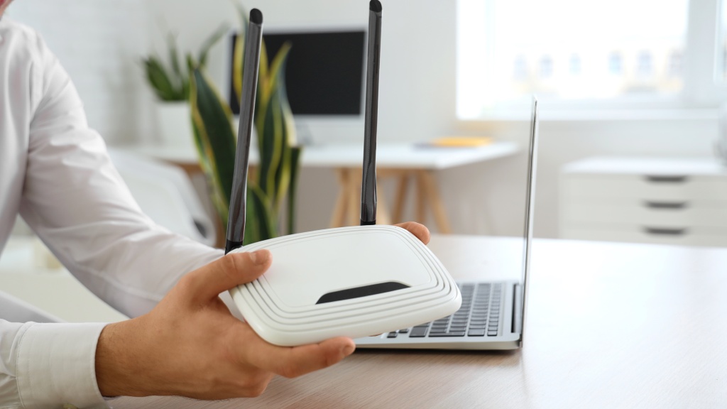 Homem segurando roteador branco e preto ao lado de notebook sobre a mesa para reposicionar o aparelho e melhorar as taxas de download e upload da internet.