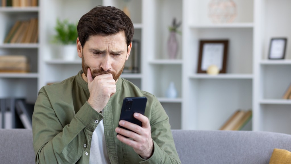 Homem de camisa jeans sorrindo, sentado à mesa de madeira com iPhone na mão, avaliando sinais para evitar golpes do WhatsApp.