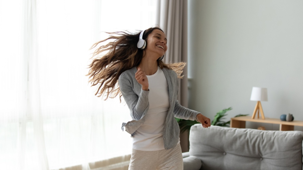Mulher jovem de camiseta branca, blusa cinza e headphones brancos, sorrindo e dançando no meio da sala com os cabelos ao vento, aproveitando boa taxa de download.