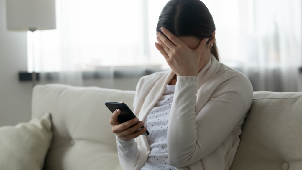 Mulher de blusa bege e camiseta branca sentada em sofá bege, segurando o celular em uma mão e cobrindo o rosto com a outra após sofrer golpe no WhatsApp.